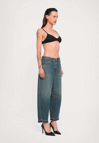 Frau in einem schwarzen Bralette, lockeren blauen Jeans und schwarzen High Heels, die seitlich steht und Tattoos auf ihren Armen und ihrem Oberkörper hat.