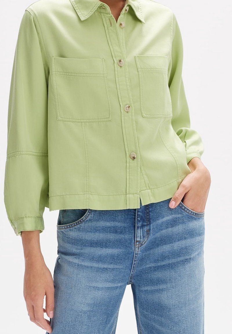 Personne portant une chemise vert clair boutonnée avec deux poches avant et un jean bleu, une main dans la poche, montrée du cou aux hanches.