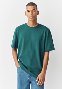 Turkio spalvos trumpomis rankovėmis medvilninė marškinėliai su apvalia iškirpte ir laisvu kirpimu. Derinami su šviesiai mėlynais džinsais.