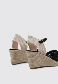 Sandales compensées avec un upper tissé noir et une bride de cheville à carreaux beiges. Plateforme en jute avec des détails texturés. Boucle ajustable.