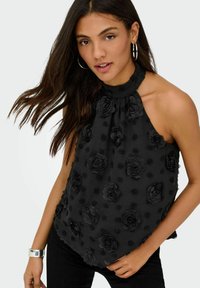 Jeune femme aux longs cheveux foncés portant un haut dos nu noir à motifs floraux en 3D et de grandes boucles d'oreilles créoles.