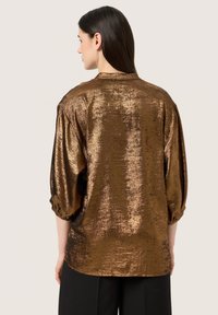 Camicia in metallo marrone con una texture lucida, maniche a sbuffo lunghe e una vestibilità ampia, caratterizzata da un collo rotondo e una leggera lucentezza.