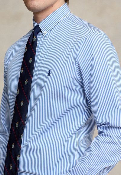 Svetlomodrá a bielo pruhovaná košeľa s priliehavým strihom, s golierom na gombíky a tmavomodrou kravatou s dekoračnými motívmi.