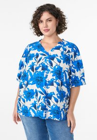 Blaue Bluse mit kurzen Ärmeln und V-Ausschnitt, die große blaue Blumen auf einem weißen Hintergrund zeigt und subtile Rüschendetails am Ausschnitt aufweist.