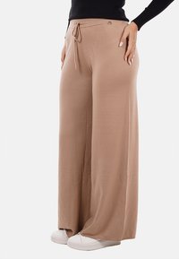 Pantaloni beige a gamba larga realizzati in tessuto morbido, con vita regolabile e tasche laterali sottili. Indossati con sneakers bianche.