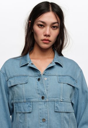 Joven con cabello largo y oscuro que lleva una chaqueta de mezclilla azul clara sobre un fondo blanco liso.