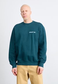 Felpa di un verde scuro con polsini e orlo a costine, caratterizzata da un piccolo logo bianco sul petto. Indossata con pantaloni beige chiaro.