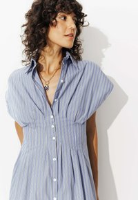 Chemise à rayures bleues, à manches courtes, avec une taille cintrée et une jupe légèrement évasée ; elle présente un col et des boutons blancs.