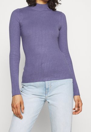 Persona que lleva un suéter morado de manga larga ajustado con textura acanalada y vaqueros azul claro, de pie contra un fondo liso.