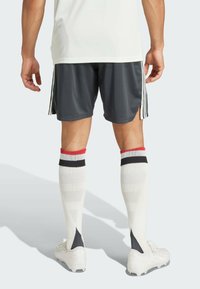 Outfit sportivo composto da una maglietta bianca, pantaloni corti grigio scuro e calze bianche al ginocchio con strisce rosse e nere, abbinate a scarpini bianchi.