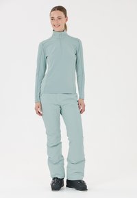 Vaaleanvihreä, pitkähihainen vetoketjullinen paita korkealla kauluksella, yhdistettynä samanvärisiin vihreisiin housuihin. Mustat hiihtokengät viimeistelevät asukokonaisuuden.