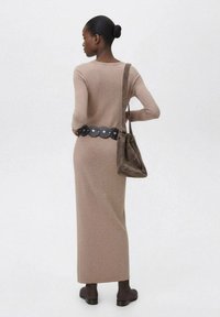 Un lungo vestito lavorato a maglia beige con vita aderente, accentuato da una cintura circolare nera. Una borsa a spalla grigia e stivaletti marroni alla caviglia completano il look.