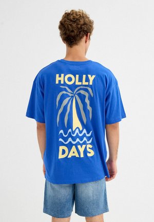 Joven con pelo rizado que lleva una camiseta azul con texto amarillo "HOLLY DAYS" y un gráfico de una palmera y olas en la espalda.