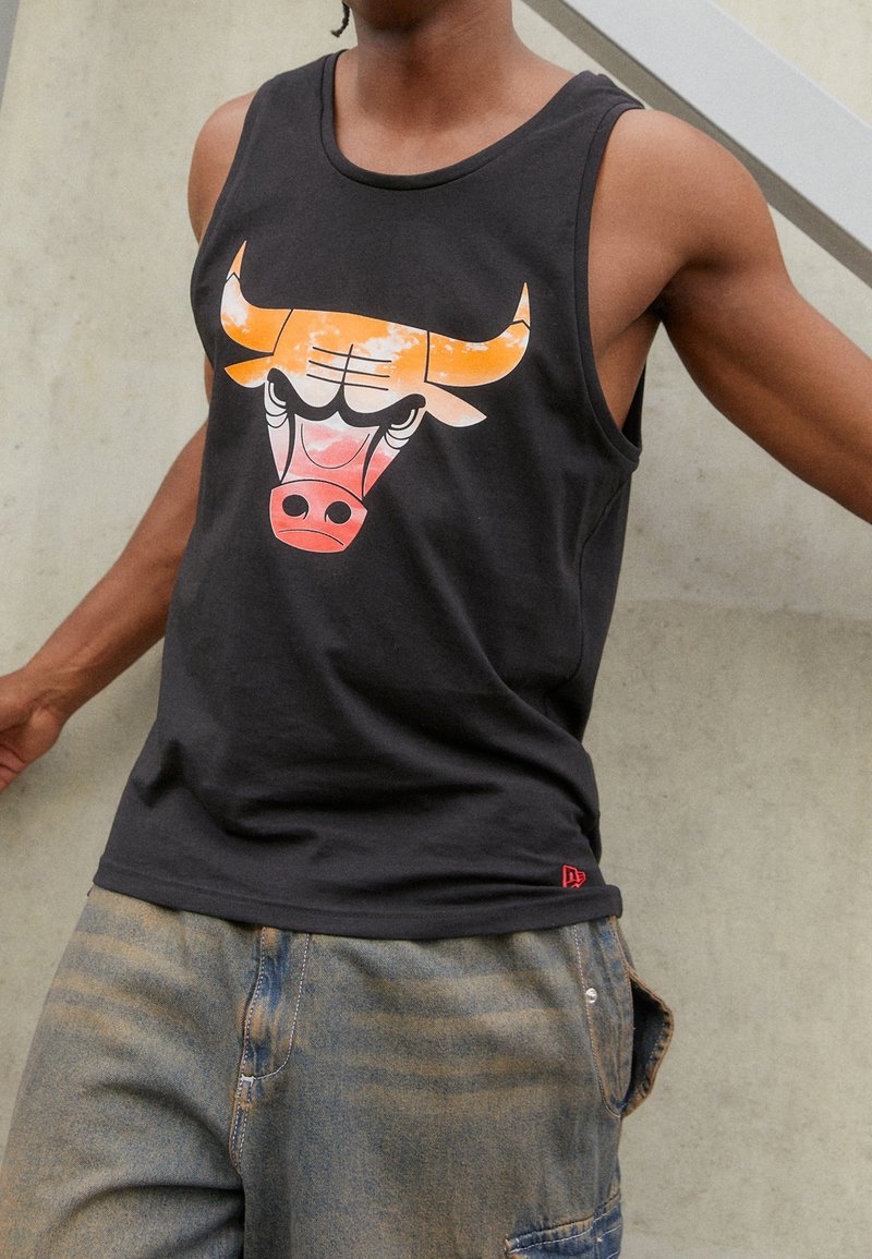 Personne portant un débardeur noir avec un logo stylisé de tête de taureau orange et rouge et un jean bleu délavé.