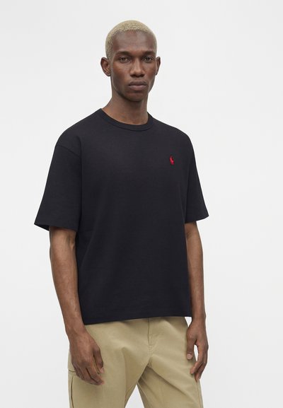 Άνδρας με κοντά ξανθά μαλλιά, φορέσει μαύρο μπλουζάκι με μικρό κόκκινο λογότυπο και μπεζ παντελόνια, στέκεται μπροστά σε απλό λευκό φόντο.