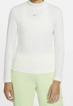 Person wearing a white long-sleeve Nike top with a turtleneck and light green pants, hands relaxed at sides.