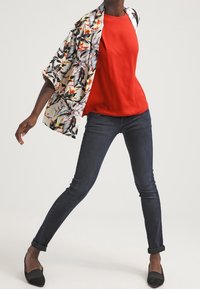 Červený top bez rukávů pod bílým a černým kimono s barevným vzorem, doplněný tmavými úzkými džínami a černými balerínami.