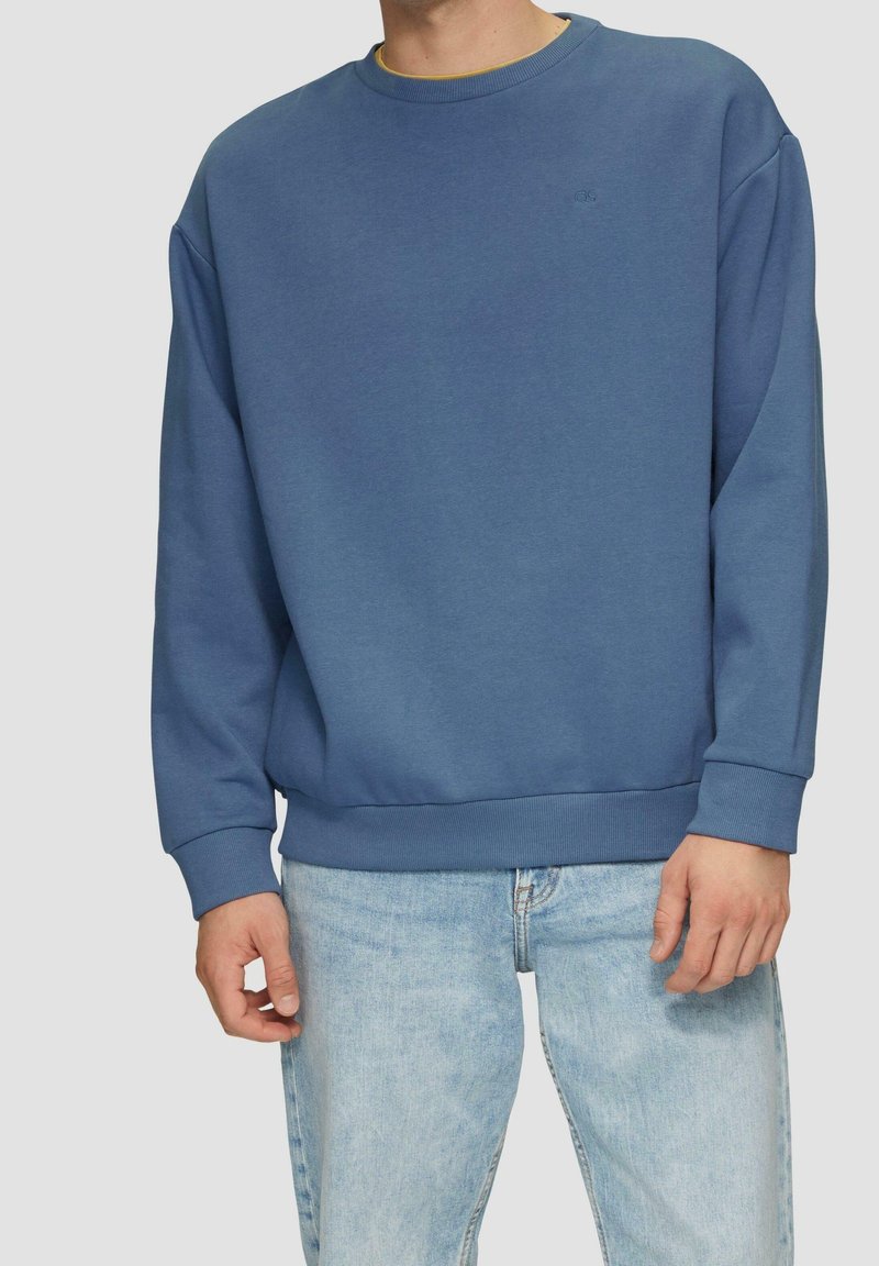 Homme portant un sweat-shirt bleu uni à col rond et un jean bleu clair, debout les bras détendus le long du corps sur un fond uni.