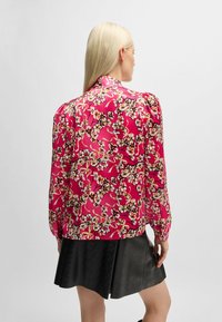 Rosa Blumenbluse mit langen Ärmeln, aus glänzendem Stoff, mit einem kunstvollen Blütenmuster und einem lockeren Schnitt, kombiniert mit einem schwarzen Rock.