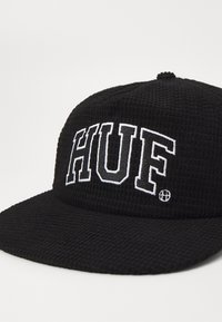 Căciulă neagră dintr-un material texturat, având un logo brodat alb "HUF" pe față. Bor vag din față cu partea superioară rotunjită.