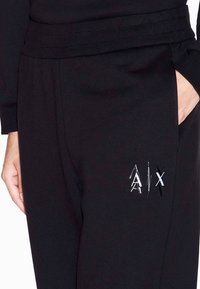 Zwarte sweatpants met een geribbelde tailleband, voorzien van een glanzend zilver en wit "A|X" logo op de linker dij en voorzakken.