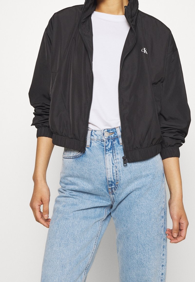 Černá lehká bomber bunda se zipem vpředu, elastickými manžetami a pasem. Stylizována s bílým tričkem a světle modrými džíny.
