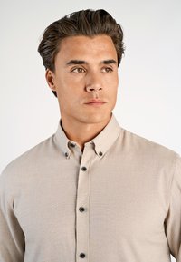 Giovane uomo con capelli scuri che indossa una camicia button-up beige, che guarda leggermente verso destra su uno sfondo chiaro e semplice.