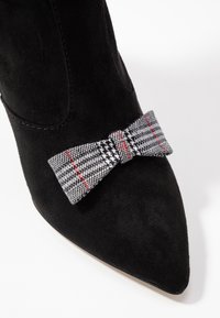Bottine en daim noir avec un bout pointu, ornée d'un nœud à carreaux gris et blanc avec des accents rouges sur le côté.