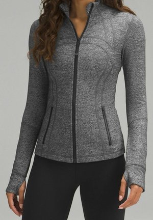 Vrouw draagt een nauwsluitend grijs sportjack met rits en duimgaten, gecombineerd met zwarte legging tegen een effen achtergrond.