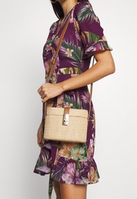 Borsa a tracolla in paglia intrecciata con forma cilindrica, tracolla e chiusura in pelle color cuoio, e texture naturale. Vestito con stampa floreale in tonalità viola.