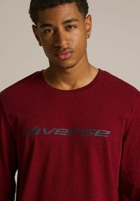 Bordowa koszula z długim rękawem, wykonana z teksturowanej tkaniny, z wyraźnym, ciemnoszarym logo "diverse" na piersi. Klasyczny krój z okrągłym dekoltem.