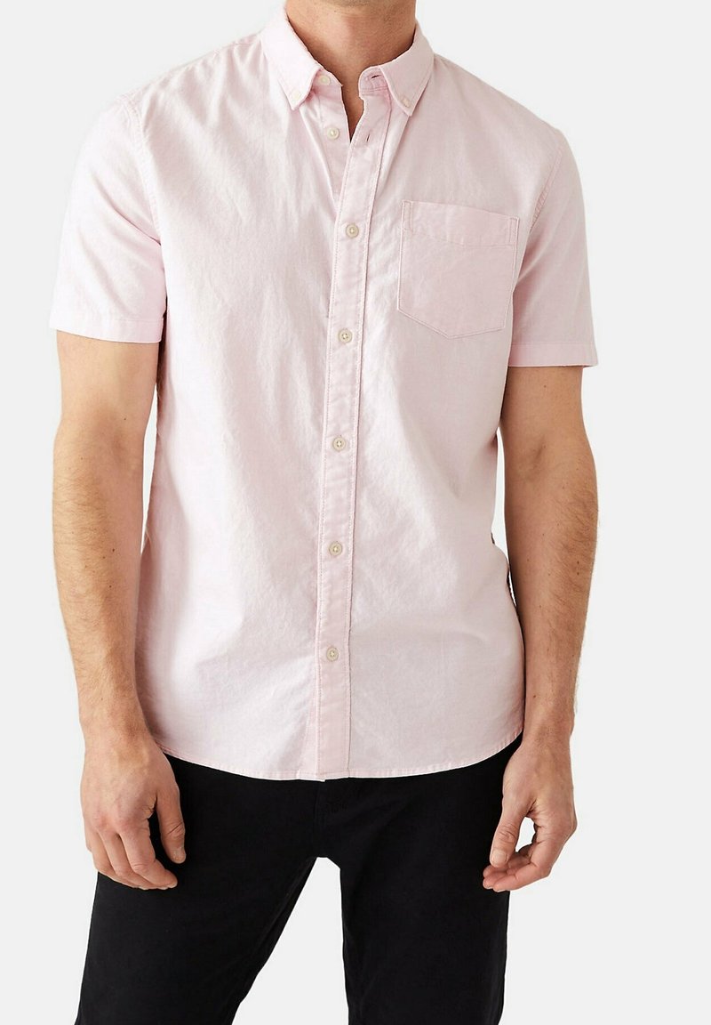 Homme portant une chemise rose clair à manches courtes avec une poche poitrine et un pantalon noir, debout devant un fond uni.