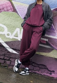 Viininpunainen verryttelypuku, väljä istuvuus, yhdistetty tumman bomber-takin kanssa. Jalkineina mustavalkoiset lenkkarit. Kaupunkimainen graffititausta.