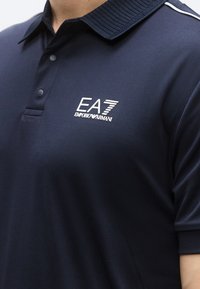 Temno modra polo majica z rebrastim ovratnikom, gumbno zaponko in belim logotipom. Tkanina z rahlim vzorcem, oprijeta zasnova in kratki rokavi.