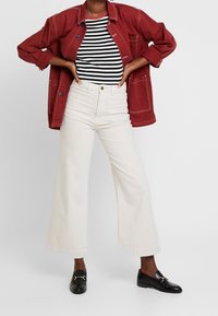 Giacca di jeans rossa con le maniche arrotolate sopra una maglietta a righe bianco e nere, abbinata a jeans bianchi a vita alta con gamba larga e mocassini neri.
