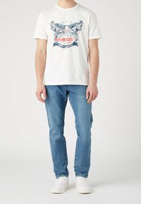 Bílý bavlněný tričko s modrým logem Wrangler a grafickým designem, kombinované se světle modrými džíny a bílými teniskami.