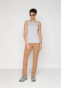 Kvinna som bär en ljusgrå ärmlös topp, bruna utomhusbyxor, grå vandringsskor och vita solglasögon, stående mot en enfärgad bakgrund.