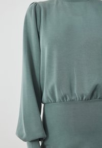 Långärmad klänning i mjukt, dämpat grönt tyg; har en insydd midja och lätt puffiga axlar för extra form och textur.