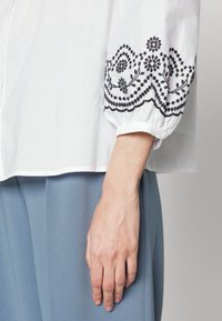 Blouse blanche avec broderie florale noire au niveau des poignets. Coupe ample avec des manches froncées. Tissu lisse, associée à un pantalon bleu clair.