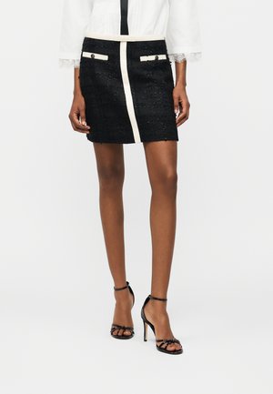 Kobieta w czarnej mini spódniczce o fakturze, z białym pionowym wykończeniem i lamówką przy kieszeniach, w parze z czarnymi sandałami na wysokim obcasie i paskami.