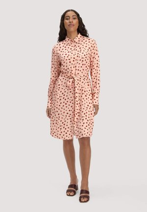 Frau steht und trägt ein hellrosa knielanges Hemdkleid mit rotem Blumenmuster, gebundenem Taillengürtel und braunen offenen Sandalen.