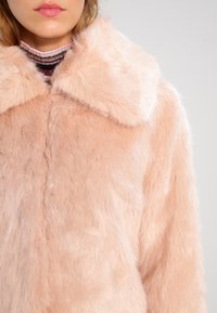 Kvinna i en mjuk, ljusrosa fuskpälsjacka med en bred krage över en randig polotröja.