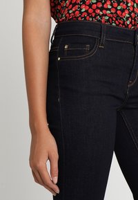 Mörkblå jeans i denim med hög midja, med guldtrådsstickning, två framfickor och en figurnära design. Närbild på sidodetaljer.