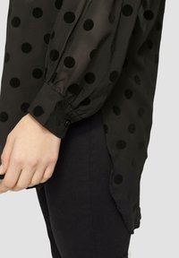 Zwarte doorschijnende blouse met gestructureerde zwarte stippen, met lange pofmouwen en een knoopmanchet, gecombineerd met donkere denim.