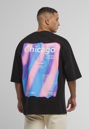 Mann sett fra siden iført svart oversized T-skjorte med fargerik "Chicago Illinois USA"-tekst og koordinater på baksiden.
