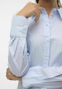 Camisa abotonada de color azul claro con un corte ajustado, mangas con pliegues y botones blancos. Textura suave y diseño de cuello clásico.