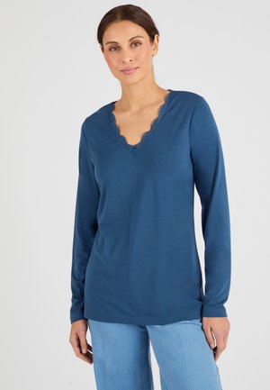 Top a maniche lunghe e scollo a V di un blu intenso con bordo in pizzo sul collo. Tessuto morbido e vestibilità rilassata. Abbinato a pantaloni azzurri chiari.