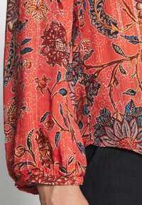 Blouse à motif floral rouge avec des manches longues et froncées. Présente des designs botaniques détaillés en bleu, noir et crème. Texture de tissu douce.