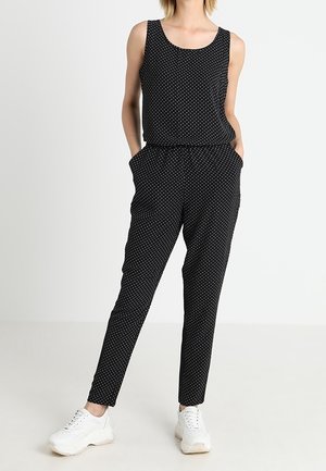 Femme portant une combinaison noire sans manches à pois, les mains dans les poches, et des baskets blanches sur un fond blanc uni.