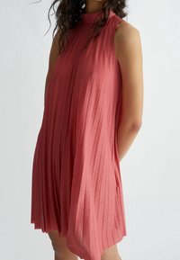 Donna che indossa un vestito senza maniche, a collo alto, plissettato e lucido di colore rosa, in piedi contro uno sfondo chiaro e uniforme.